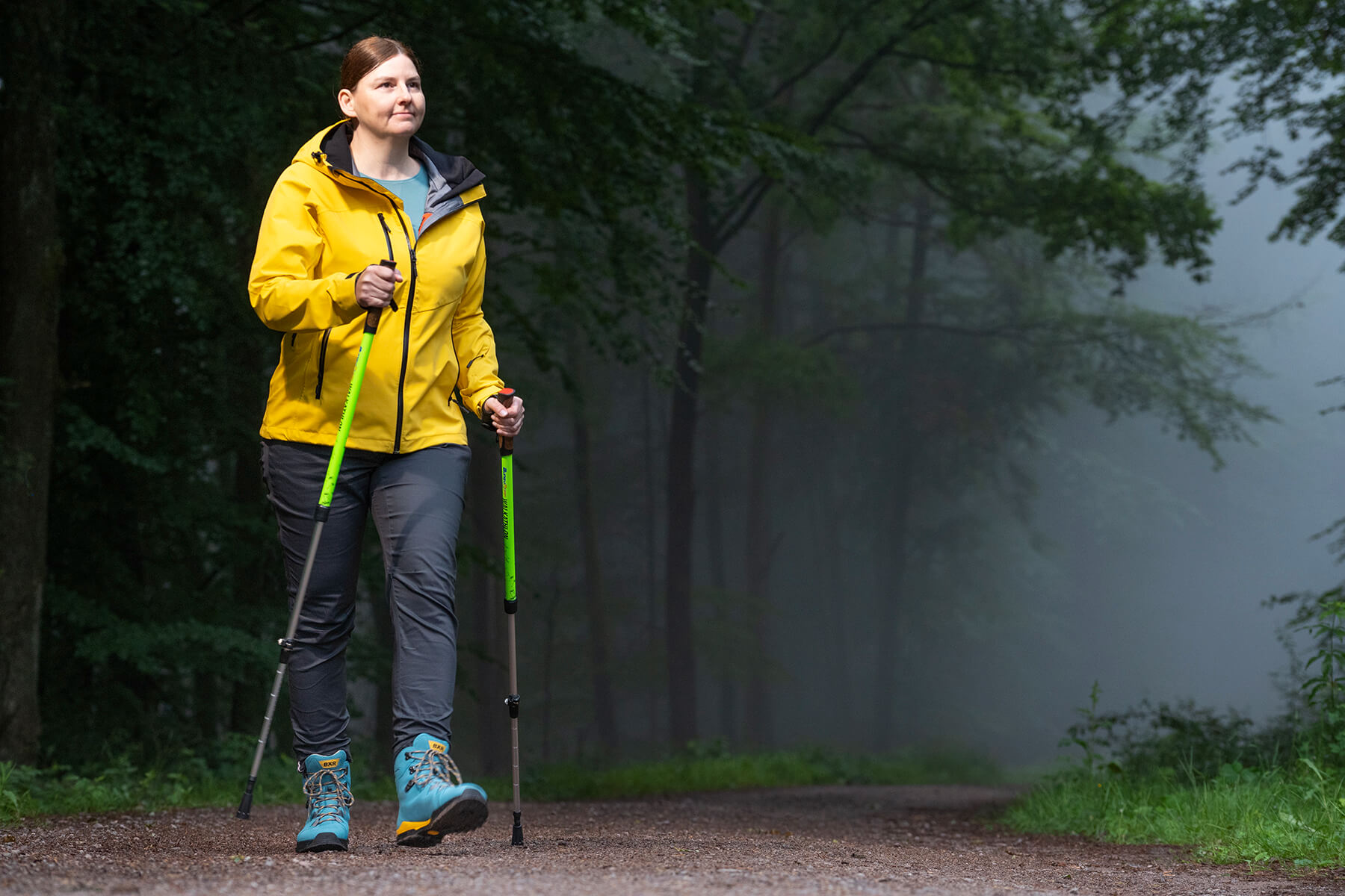 Frau mit gelber Jacke und grünen Trekkingstöcken beim Nordic Walking im nebligen Wald