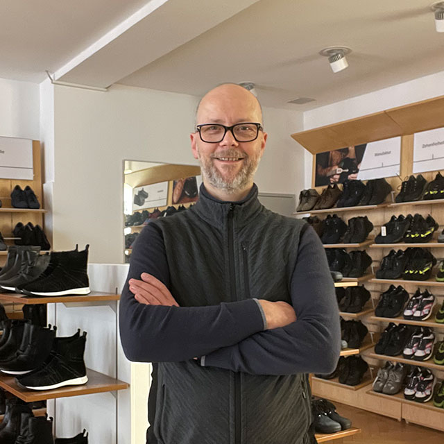 Ein lächelnder Mann steht mit verschränkten Armen in einem Schuhgeschäft. Im Hintergrund sind Regale mit verschiedenen Sportschuhen zu sehen.