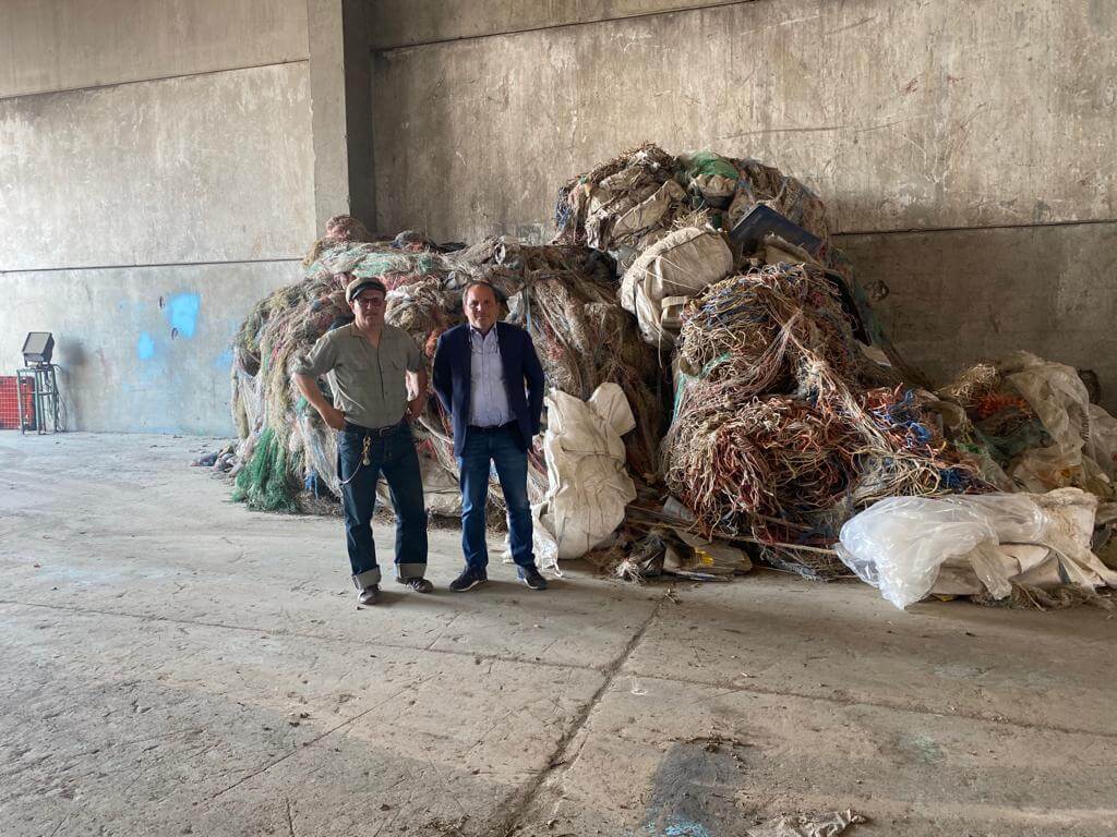 Zwei Männer stehen in einem Lagerhaus vor einem großen Haufen recyceltem Material aus Kunststoff und Metall.