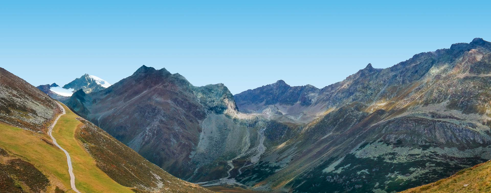 Berglandschaft in den Alpen mit einem Pfad, der durch grüne und felsige Hügel führt, vor einem klaren blauen Himmel