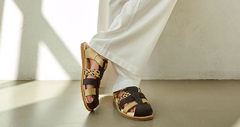 Damenfüße in stilvollen, braun-beigen Ledersandalen, weiße Hose, modernes Design