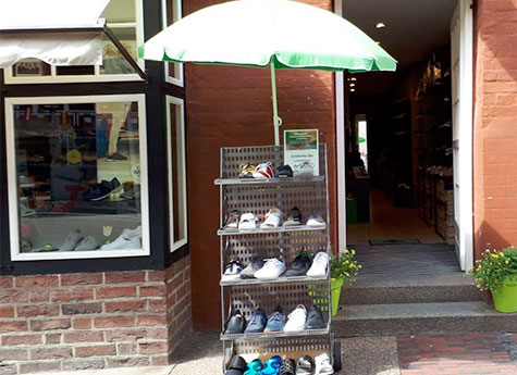 Ein Schuhständer vor einem Geschäftseingang, geschützt von einem grünen Sonnenschirm, mit verschiedenen Sportschuhen ausgestellt.