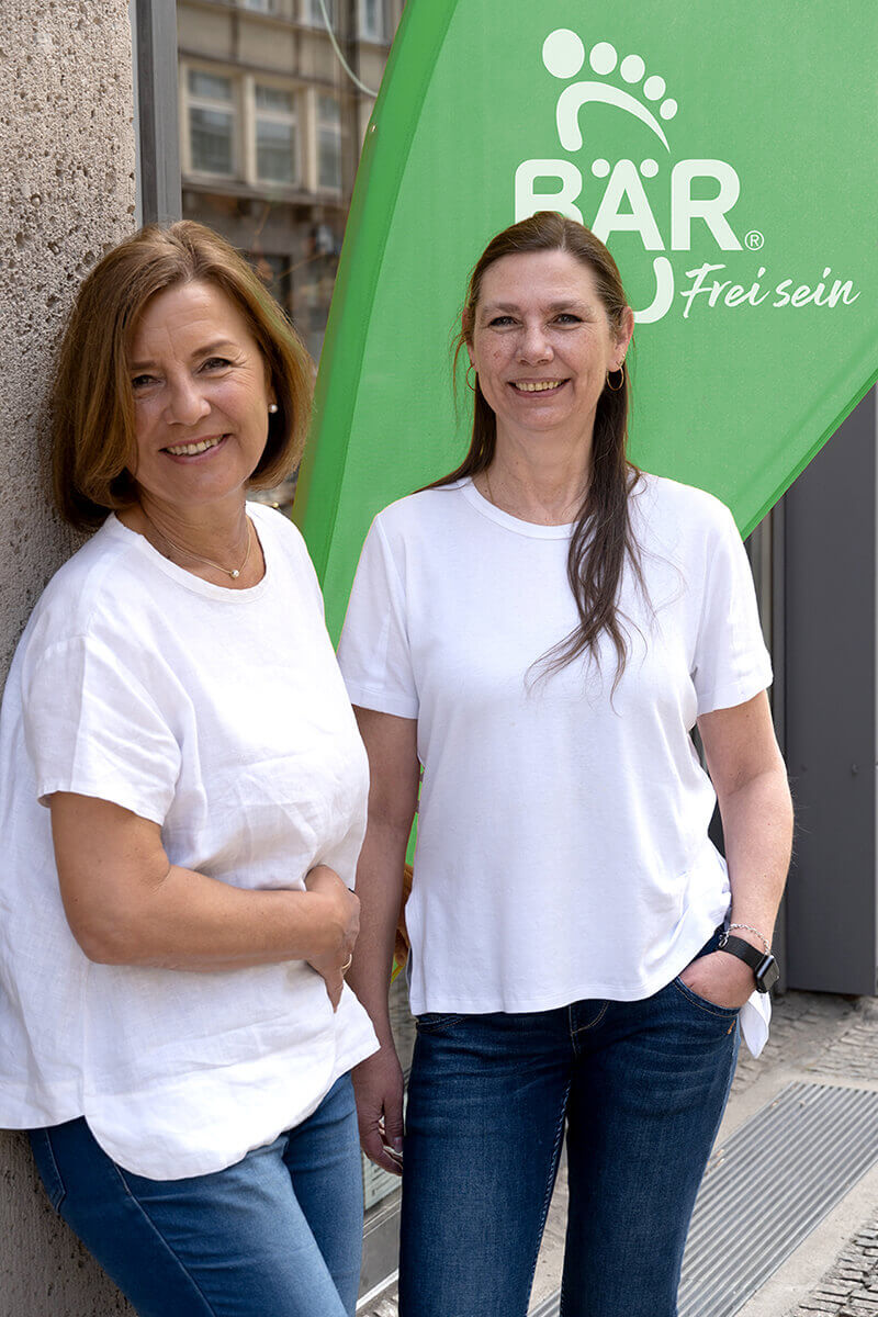 Zwei lächelnde Frauen in weißen T-Shirts und Jeans vor einem grünen Banner mit BÄR-Logo.