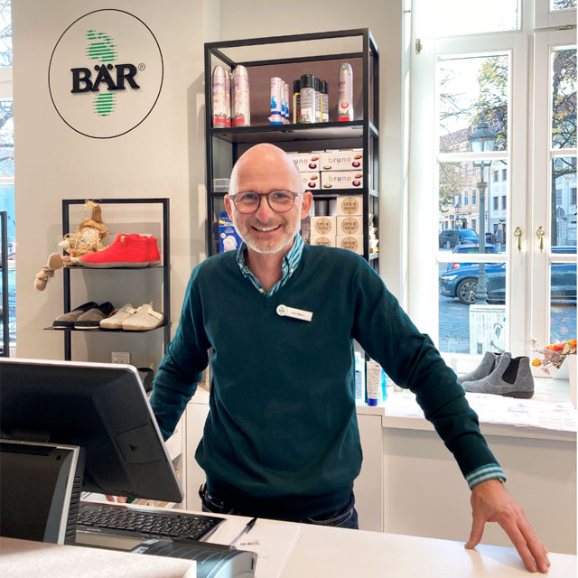 Ein freundlicher Mitarbeiter steht lächelnd hinter der Theke in einem Schuhgeschäft, umgeben von Schuhen und Verkaufsartikeln. Im Hintergrund sind Regale und Produkte sichtbar.