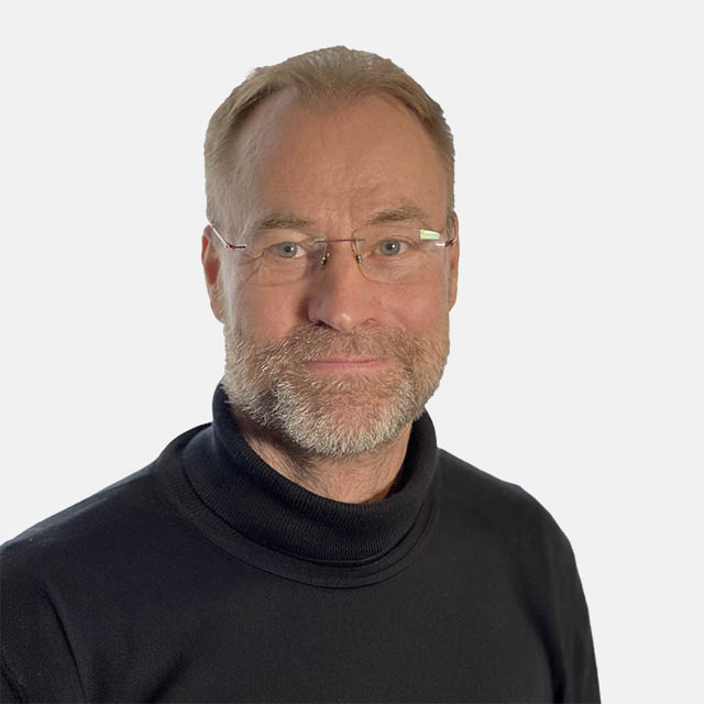 Ein lächelnder Mann mittleren Alters mit Brille und Bart, trägt einen schwarzen Rollkragenpullover vor einem neutralen Hintergrund