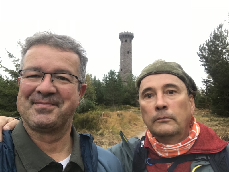 Zwei Männer beim Wandern vor einem alten Steinturm in einem bewaldeten Gebiet
