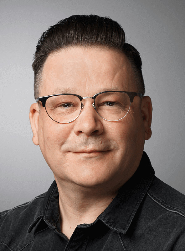 Mann mit Brille, kurzem dunklem Haar und dunklem Hemd lächelt in die Kamera