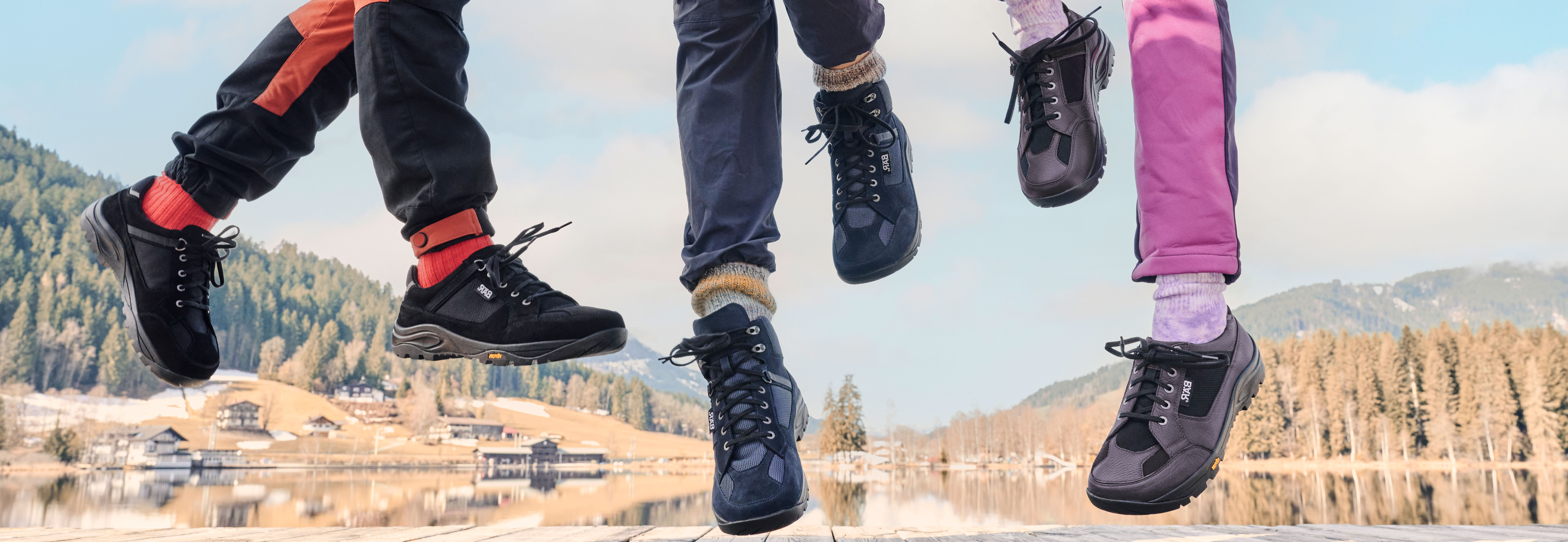 Füße in Outdoor-Schuhen springen in die Luft, vor einer malerischen Landschaft mit Bergen und einem See im Hintergrund.