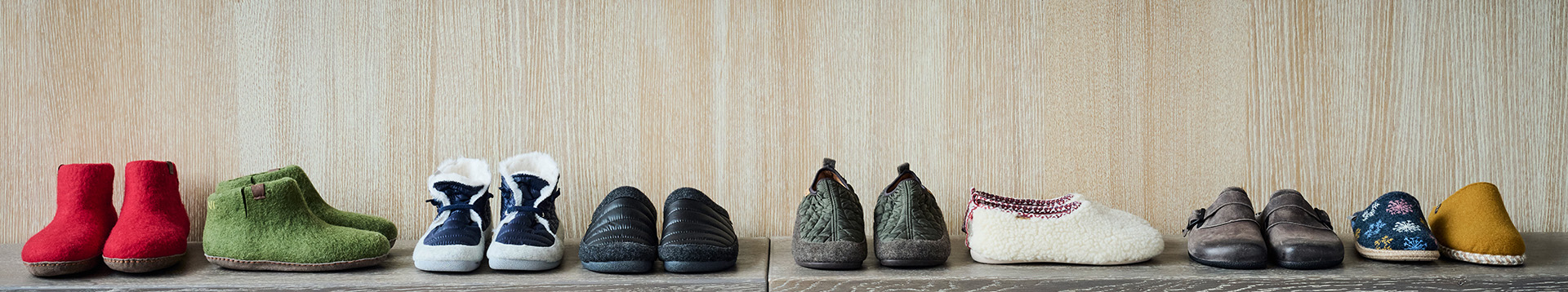 Eine Auswahl an Hausschuhen in verschiedenen Farben und Designs vor einer Holzwand auf einem rustikalen Regal