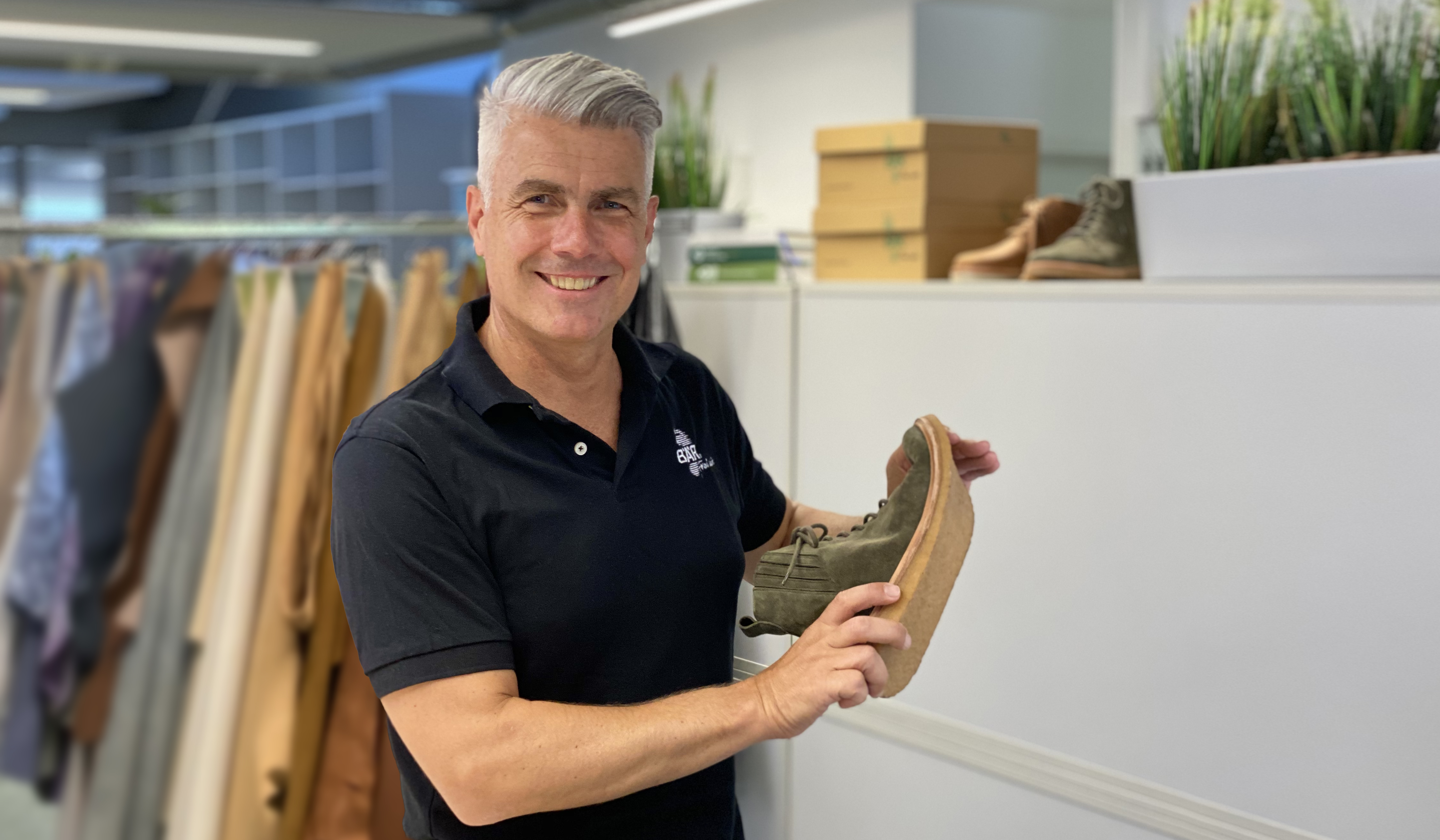 Mann mit grauen Haaren in schwarzem Polohemd hält grünen Schuh im Büro mit Kleiderständern im Hintergrund