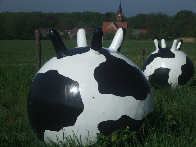 Zwei große, kugelförmige Skulpturen in schwarz-weißer Kuhoptik auf einer grünen Wiese vor einem Dorf mit Kirchturm.