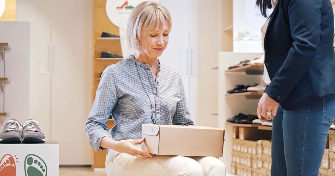 Frau sitzt mit Schuhkarton in einem Schuhgeschäft, im Hintergrund Regale mit Schuhen.
