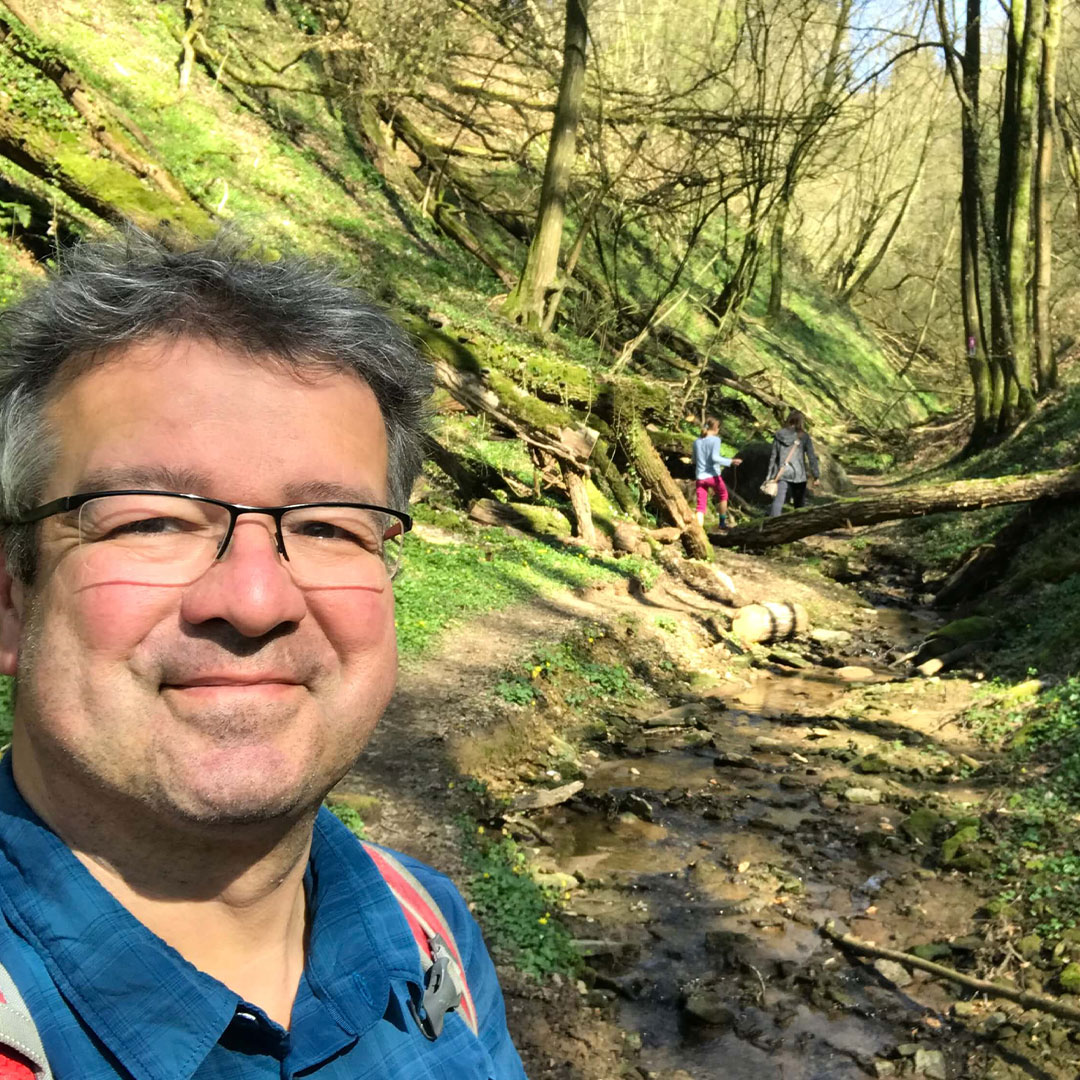 Ein Mann mit Brille und Rucksack steht auf einem schattigen Waldweg neben einem kleinen Bach, weitere Wanderer sind im Hintergrund zu sehen.