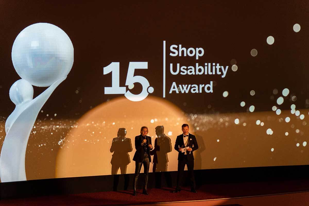 Moderatoren stehen auf der Bühne bei der 15. Verleihung des Shop Usability Awards, mit Award-Logo im Hintergrund.