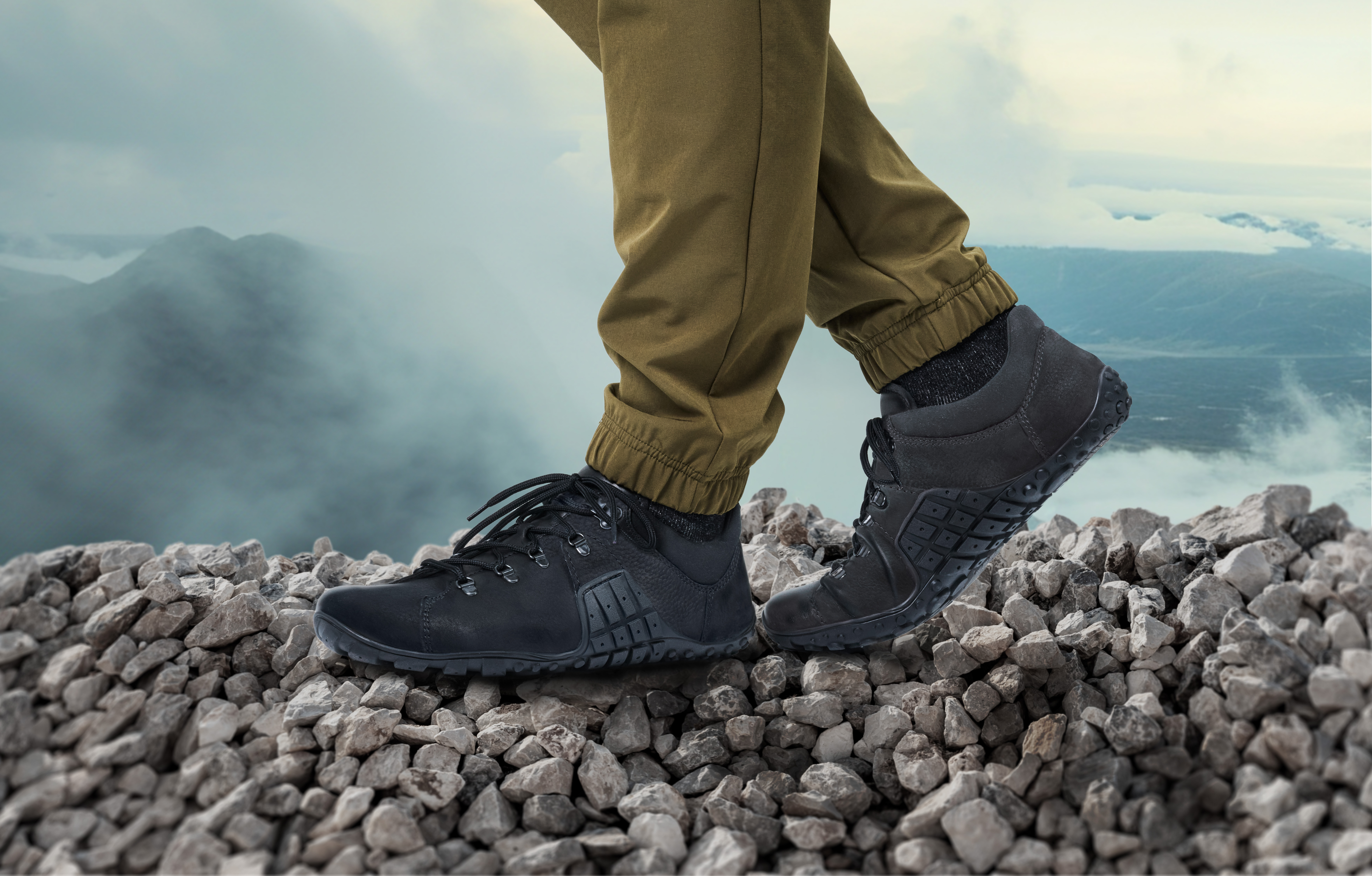 Person in Outdoor-Hosen trägt schwarze Wanderschuhe und steht auf einem steinigen Pfad mit Bergen im Hintergrund