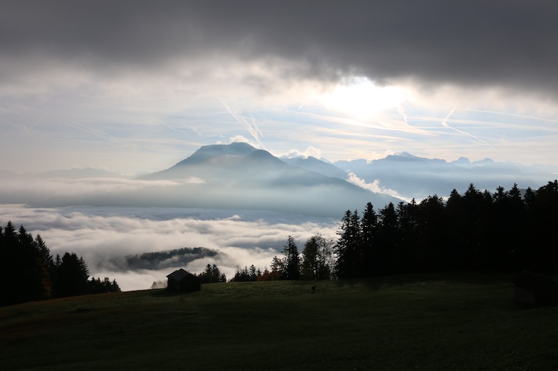 Ein malerischer Blick auf eine Berglandschaft mit dichter Wolkendecke und aufgehender Sonne, umgeben von Wäldern und Wiesen.