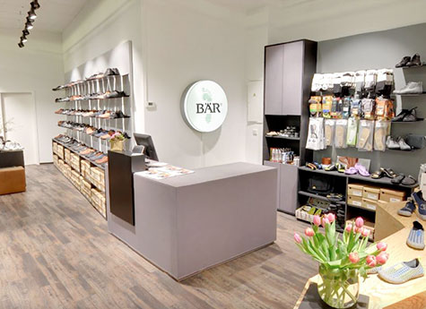 Innenansicht eines modernen Schuhladens mit Regalen voller Schuhe und Accessoires, einem grauen Tresen und einem Blumenstrauß auf einem Tisch