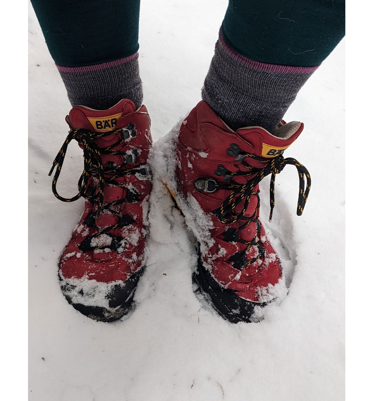 Rote Wanderschuhe mit Schnee bedeckt, getragen von einer Person mit grauen Socken auf verschneitem Boden.
