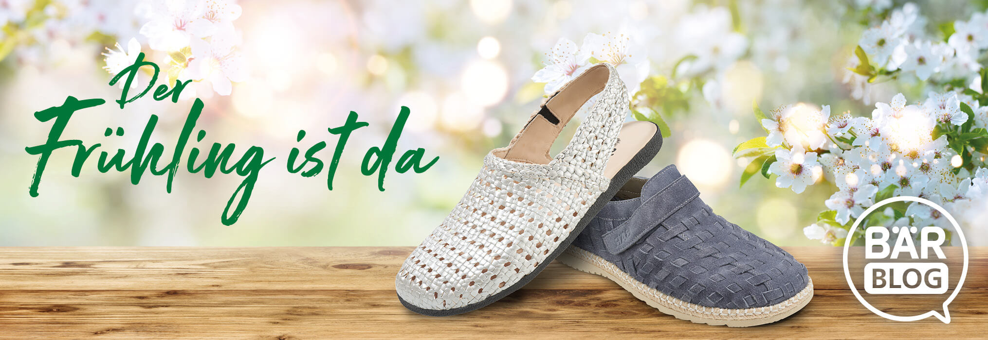 Zwei gewebte Frühlingsschuhe aus hellem und dunkelgrauem Material auf einem Holztisch, im Hintergrund blühende Blumen. Text: Der Frühling ist da, Bär Blog.
