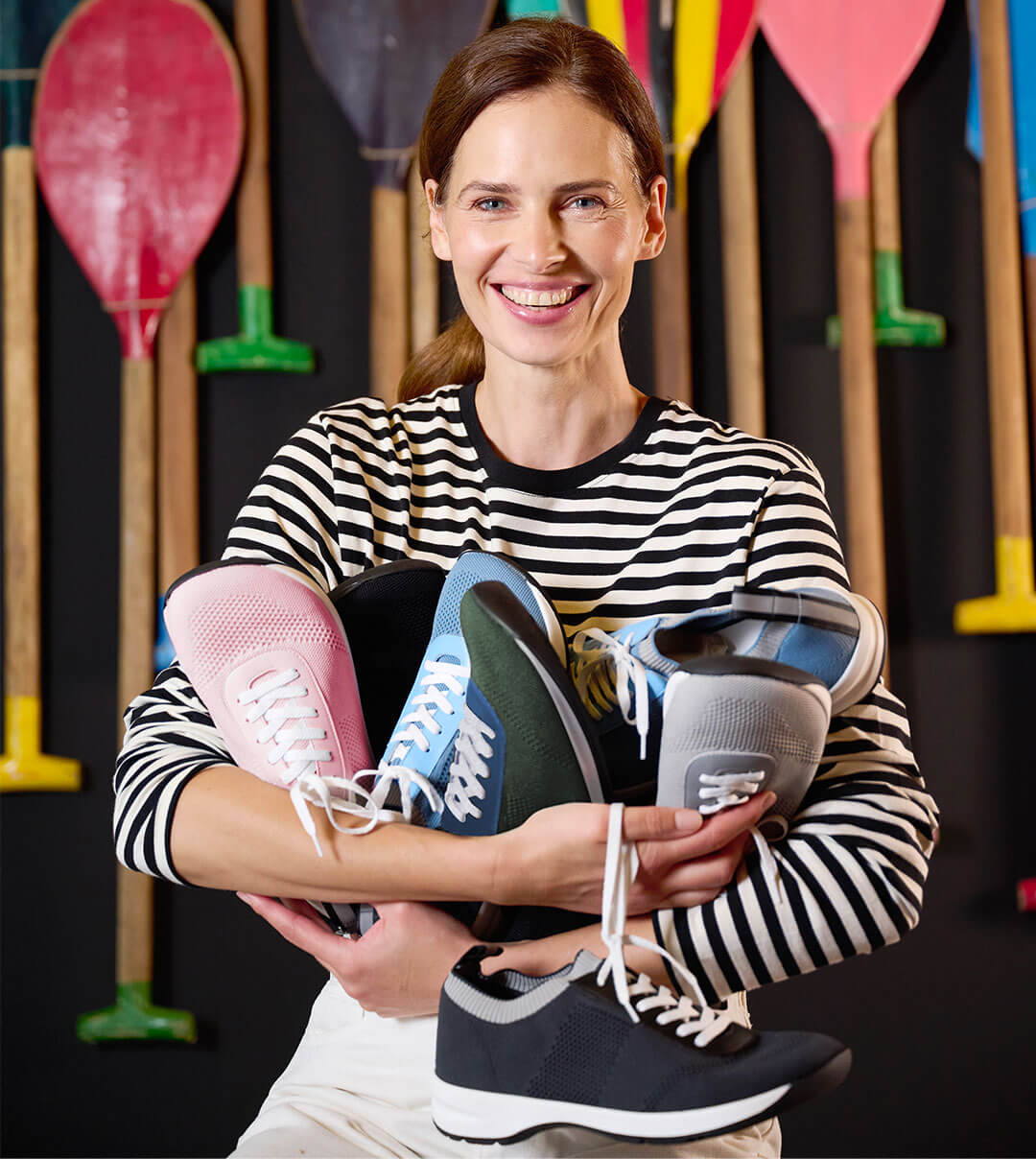 Frau in gestreiftem Shirt hält verschiedene bunte Sneaker vor Paddelhintergrund