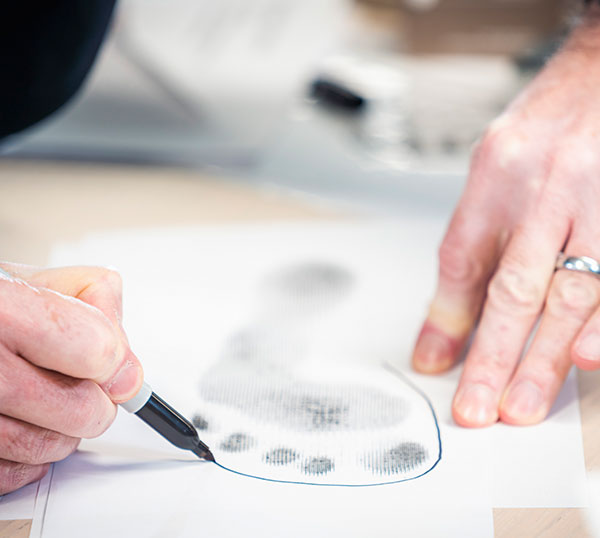 Nahaufnahme einer Person, die mit einem Stift den Umriss eines Fußabdrucks auf einem Blatt Papier zeichnet