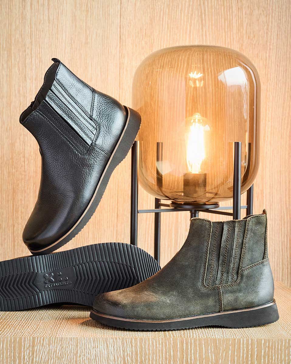 Schwarze und braune Chelsea Boots neben einer stilvollen Lampe auf Holzoberfläche