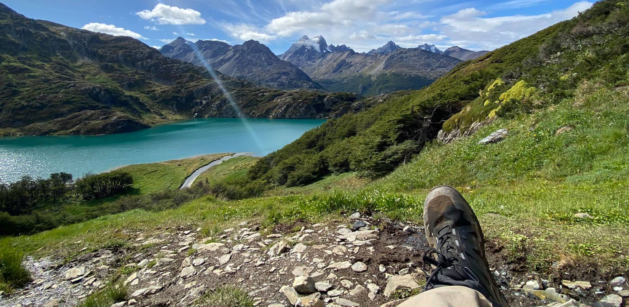 Person entspannt sich mit Wanderschuhen am Seeufer, umgeben von Bergen und blauem Himmel