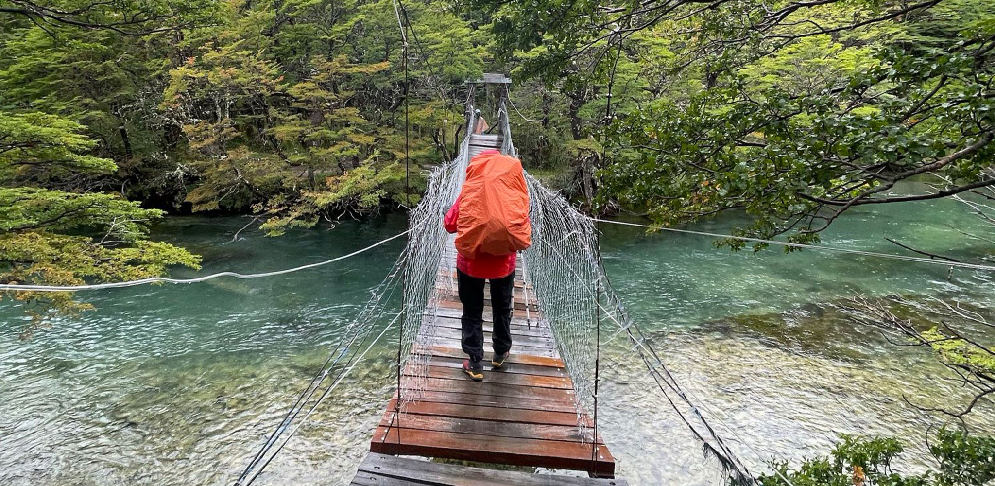 Person mit orangem Rucksack überquert eine Hängebrücke über einen klaren Fluss, umgeben von grünem Wald.