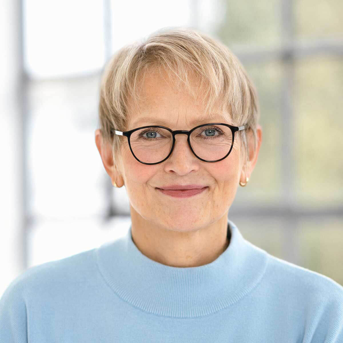 Porträt einer älteren Frau mit Brille und blauem Pullover in einem hellen Raum