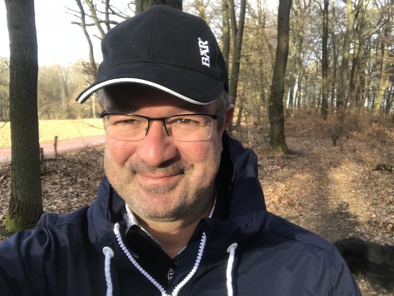 Ein Mann mit Brille und schwarzer Kappe steht lächelnd in einem sonnigen Wald.