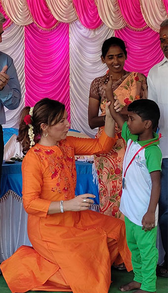 Frau in orangefarbener traditioneller Kleidung kniet und begrüßt ein Kind mit Medaille bei Feier, während andere Erwachsene zusehen